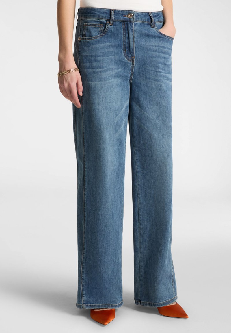 Jean en denim taille large de couleur moyenne, doté d'un design classique à cinq poches, d'une fermeture à bouton et d'un ourlet effiloché, associé à des talons aiguilles orange.