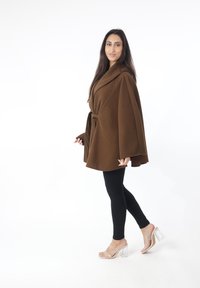 Capa marrón de gran tamaño con cinturón en la cintura, mangas anchas y tela texturizada, combinada con leggings negros y sandalias de tacón transparente.