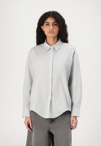 Camicia a maniche lunghe a righe grigia chiara con colletto a punta, pattina con bottoni e una tasca sul petto. Presenta una vestibilità rilassata e oversize.