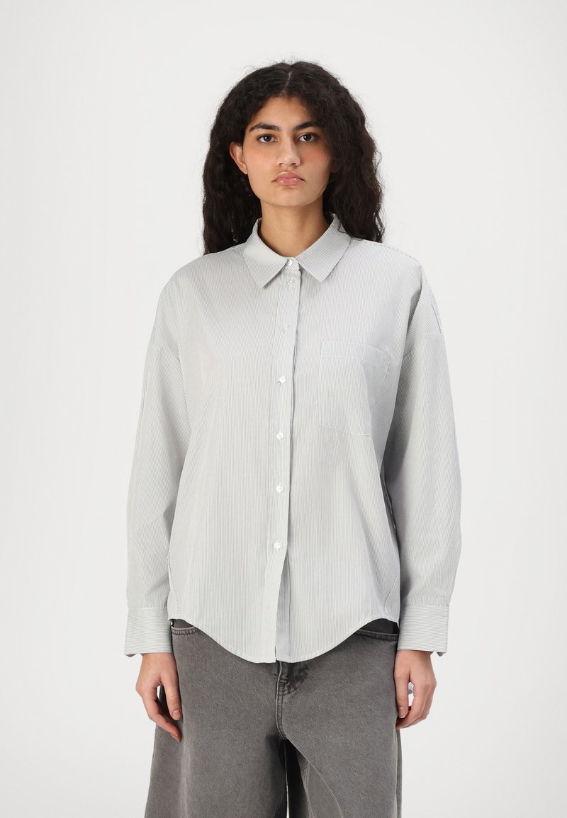 Camicia a maniche lunghe a righe grigia chiara con colletto a punta, pattina con bottoni e una tasca sul petto. Presenta una vestibilità rilassata e oversize.