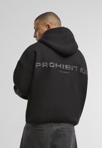 Schwarzer Kapuzenpullover mit großem, erhabenem "PROHIBIERT"-Text auf dem Rücken. Der Stoff wirkt weich und hat eine lässige Passform.