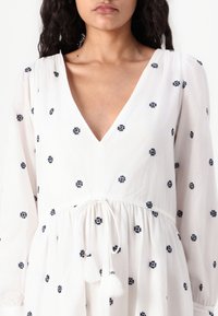 Vit klänning med marinblå broderade blommönster, V-ringning, långa ärmar, dragsko i midjan med tofsar, lätt, texturerad tyg.