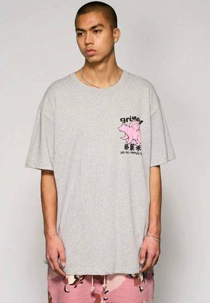 Jonge man draagt een oversized lichtgrijs T-shirt met een roze berenafbeelding en zwarte tekst, gecombineerd met roze en bruine camouflage shorts.