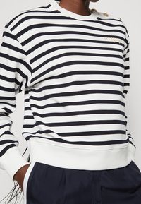 Maglione a righe con strisce orizzontali blu navy e bianche, polsini a costine e texture morbida. Decorato con un accento logo dorato.