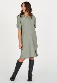 Olivgroene overhemdjurk met een knoopsluiting aan de voorkant, korte opgerolde mouwen en een losse pasvorm. Draagt daarbij kniehoge zwarte leren laarzen.