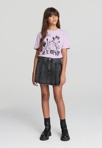 T-shirt grafico lilla con illustrazioni nere, abbinato a una gonna di jeans nera a vita alta e stivali neri con tacco chunky.