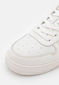 Vit lädersneaker med rundad tå, perforerad ovandel och platta vita snören. Sulan är texturerad och något upphöjd.