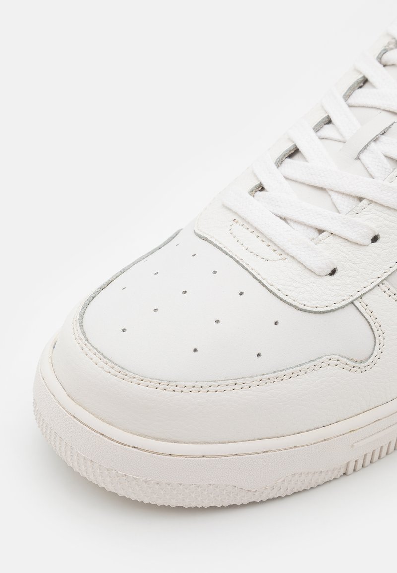 Zapatilla de cuero blanca con punta redondeada, parte superior perforada y cordones blancos planos. La suela es texturizada y ligeramente elevada.