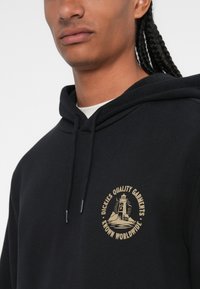 Hoodie preto com um logotipo redondo em dourado, apresentando um design de farol e o texto "Dickies Quality Garments Known Worldwide". Material de algodão.