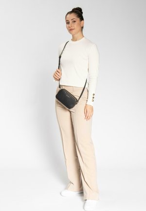 Bolso cruzado negro texturizado con una correa desmontable, combinado con una blusa de manga larga crema y pantalones de campana beige en un entorno neutro.
