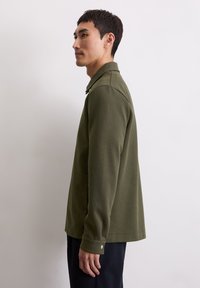 Oliwkowa, długirękawowa koszulka polo z kołnierzykiem i guzikami. Miękka, teksturowana tkanina, luźny krój, rozcięcia po bokach u dołu.