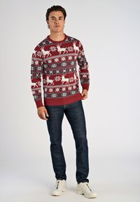 Roter Strickpullover mit weißen Rentier- und Schneeflockenmuster, Rundhalsausschnitt, gerippte Bündchen und Saum, kombiniert mit dunklen Jeans und weißen Sneakers.