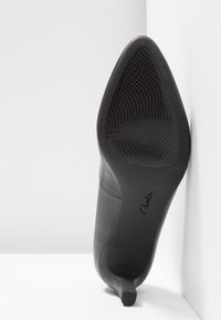 Chaussure à talon haut en cuir noir, bout pointu, texture lisse, avec une semelle en caoutchouc dotée d'une prise antidérapante à motif. Logo visible sur la semelle.