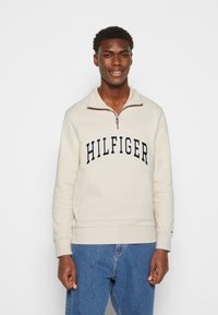 Felpa beige con zip e logo "HILFIGER" in blu navy, tasca a marsupio e polsini a costine. Abbinarla a jeans denim.