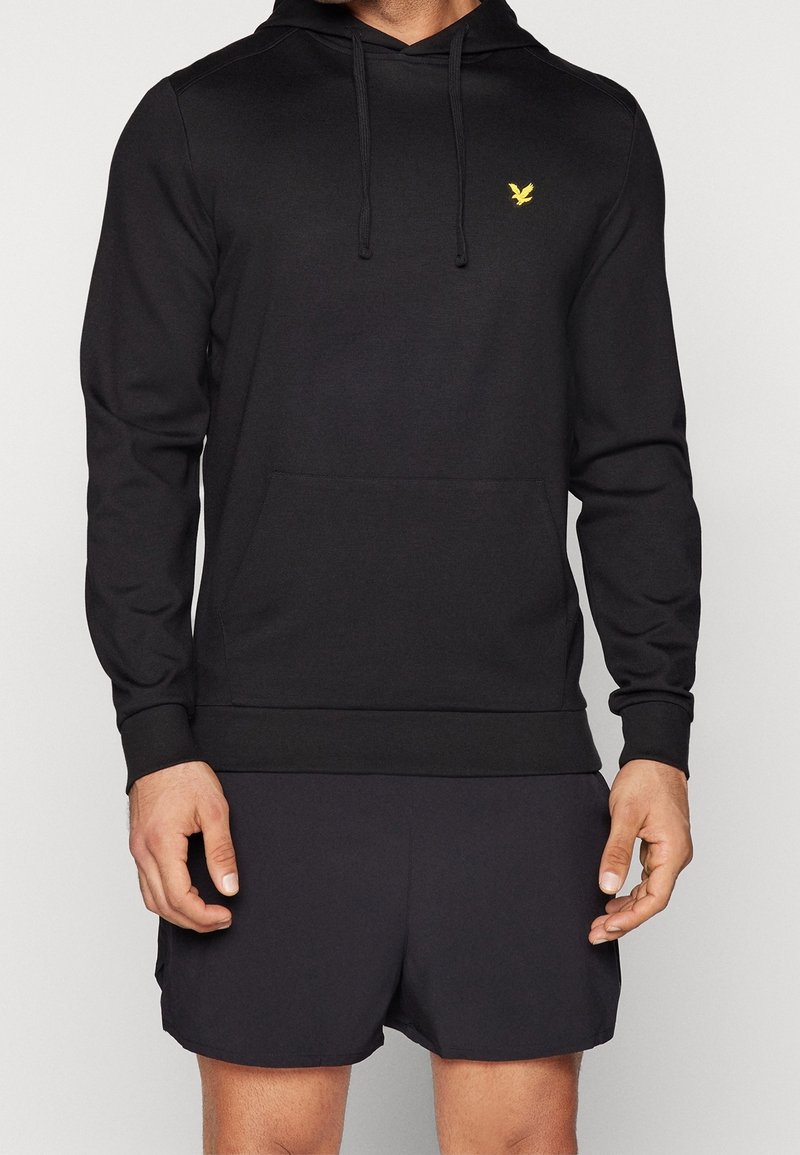 Homme portant un sweat à capuche noir avec un logo d'oiseau jaune sur la poitrine et un short noir, visible du cou au milieu de la cuisse, sur un fond uni.