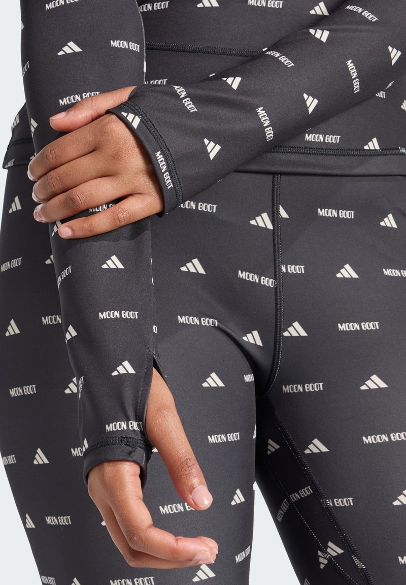 Schwarzes Sportoberteil mit tailliertem Design, das ein wiederholtes Muster aus dem weißen Text und Logo "MOON BOOT" aufweist, lange Ärmel und Daumenlöcher an den Bündchen hat.