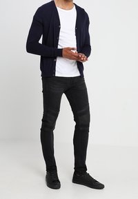 Cárdigan navy sobre una camiseta blanca, combinado con jeans negros con acentos de cuero. Modelo de pie en zapatillas negras contra un fondo claro.