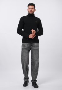 Suéter negro de punto con cuello alto y superficie texturizada, combinado con jeans grises de pierna recta y zapatillas negras. Detalle de acento amarillo sencillo.