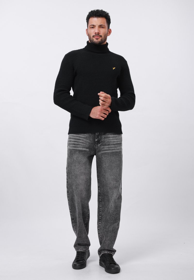 Maglione a collo alto nero in maglia con superficie testurizzata, abbinato a jeans dritti grigi e scarpe da ginnastica nere. Dettaglio accentato semplice in giallo.