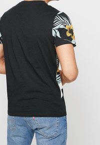 Muž v černém tričku s rukávy s modrožlutým květinovým vzorem a světle modrých džínách, pohledem zezadu.