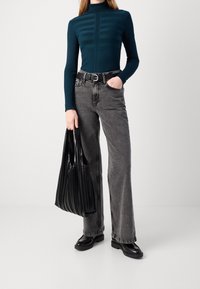 Haut en turtleneck côtelé teal avec une texture rayée, associé à un jean en denim gris taille haute et un sac à main plissé noir. Bottines chunky noires.