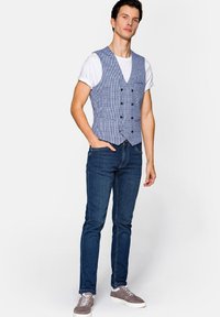 Niebieska i biała kamizelka w kratę z dekoltem w kształcie V, zapinana na podwójne guziki, noszona na białym T-shircie, w zestawie z ciemnymi dżinsami i szarymi trampkami.