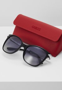 Svart oversized solbriller med gradientlinser. Teksturert rødt etui med preget "GUESS"-logo. Vinklet design med sølvfargede detaljer.