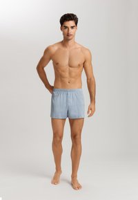 Světle modré bavlněné trenýrky s elastickým pasem, single button fly a hladkou texturou, vystavené na modelovi bez trička.