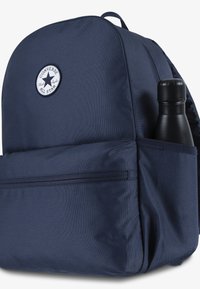 Sac à dos bleu marine avec un logo Converse All Star rond, en tissu texturé, poche avant et poche latérale tenant une bouteille noire.