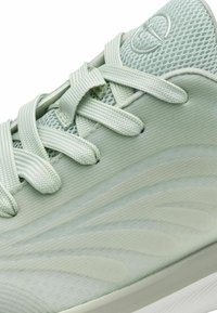 Chaussure de sport vert clair dotée d'une tige en mesh texturé, de lacets plats et de motifs ondulés sur le côté. Semelle souple pour un amorti.