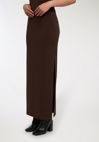 Abito maxi attillato marrone con spacchi laterali, realizzato in tessuto morbido. Abbinato a stivali alla caviglia neri con tacco, che mettono in evidenza un design affusolato.