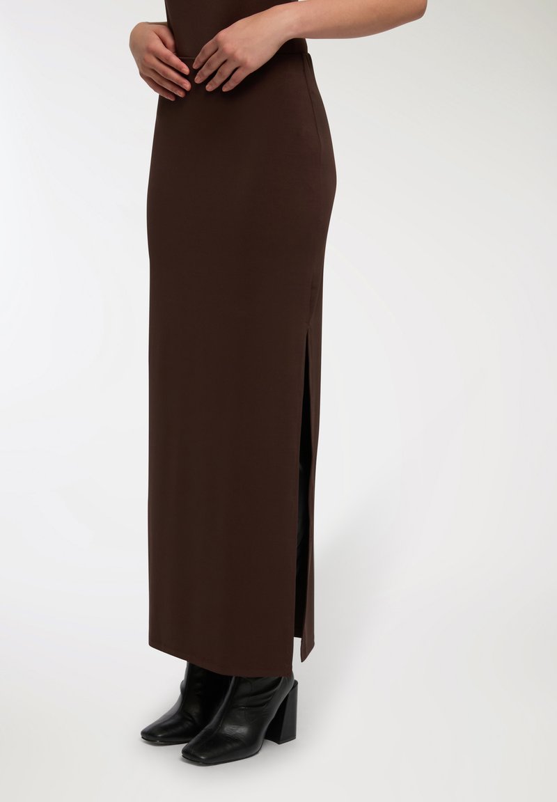 Abito maxi attillato marrone con spacchi laterali, realizzato in tessuto morbido. Abbinato a stivali alla caviglia neri con tacco, che mettono in evidenza un design affusolato.