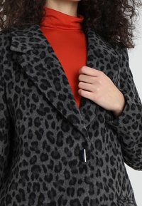 Šedý béžový kabát s leopardím vzorem, s klopovým límcem, zapínáním na jeden knoflík a měkkou texturou, nošený přes jasně červený rolák.