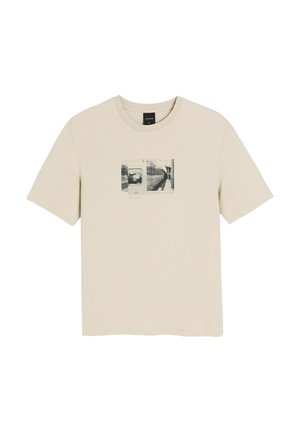 Beżowa koszulka z krótkim rękawem z czarno-białą grafiką przedstawiającą pociąg metra i peron, z napisem "Diverse System" na środku przodu.