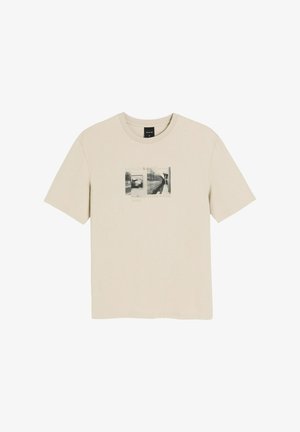 Beżowa koszulka z krótkim rękawem z czarno-białą grafiką przedstawiającą pociąg metra i peron, z napisem "Diverse System" na środku przodu.