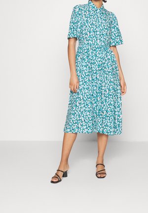 Kobieta ubrana w miętową sukienkę midi z białym kwiatowym wzorem, krótkimi bufkami, kołnierzykiem i czarnymi sandałami na obcasie na prostym tle.