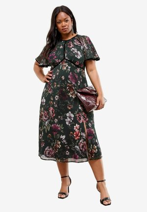 Blumen-Midi-Kleid mit kurzen, flatternden Ärmeln, auf einem dunkelgrünen Hintergrund mit rosa und weißen Blumen sowie einem seitlichen Schlitz. Kombiniert mit einer burgunderfarbenen Tasche.