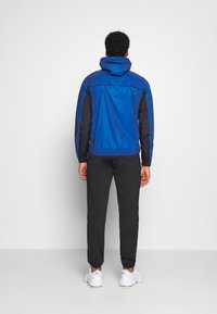 Lightweight blue and black windbreaker jacket with a hood, elastic cuffs, and a simple design, paired with black jogger pants and white shoes.