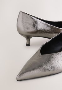 Fémes ezüst anyagból készült, hegyes orrú magassarkú cipő, amely texturált felülettel és vékony, rövid sarokkal rendelkezik. Nyitott oldalak fekete belsőt tárnak fel.