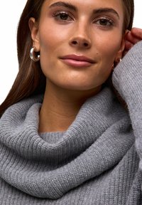 Maglione grigio a coste con scollo a cappuccio. La modella ha i capelli castani di lunghezza media e indossa orecchini a cerchio in argento. Tessitura della pelle liscia.
