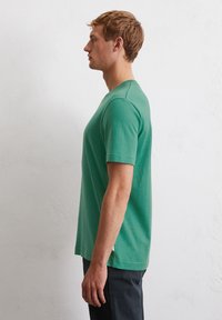T-shirt vert à manches courtes fabriqué en tissu doux, avec un col rond et une coupe décontractée. Étiquette de couture latérale visible près de l'ourlet gauche.