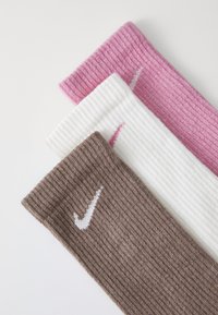 Tři páry žebrovaných ponožek v hnědé, bílé a růžové barvě, přičemž každý pár má kontrastní bílé logo Nike.
