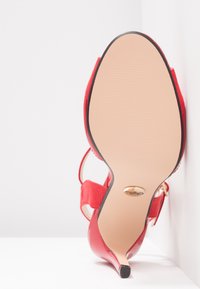 Chaussure à talons hauts rouge avec bout pointu et bride à la cheville, semelle tournée vers l'avant sur un fond blanc à angles.