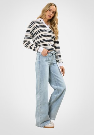 Donna con lunghi capelli biondi che indossa una maglia a maniche lunghe a righe bianche e nere, jeans larghi e chiari, e scarpe da ginnastica bianche.