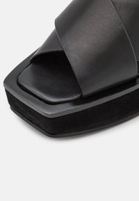 Sandale plateforme noire avec large sangle en cuir, bout carré ouvert et semelle épaisse recouverte de suède sur fond blanc uni.
