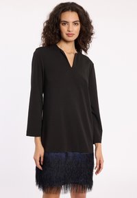 Schwarzes Tunika-Kleid mit V-Ausschnitt und langen Ärmeln, das einen strukturierten, fransigen Saum in Dunkelblau aufweist und einen kontrastierenden Schichteffekt erzeugt.
