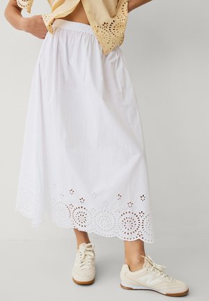 Kobieta ubrana w białą midi spódnicę z kwiatowym oczkowym obszyciem, beżowy top z krótkim rękawem i oczkami oraz beżowe sneakersy na sznurowadła na jednolitym tle.