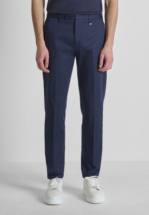 Hombre vistiendo pantalones ajustados azul marino y zapatillas blancas, de pie frente a un fondo liso de color gris claro.