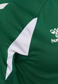 Zielona sportowa koszulka z białym paskiem w kształcie litery V i logo Hummel na piersi, ukazująca szczegółowe przeszycia i fakturę tkaniny.