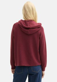 Burgundijas krāsas kapuce ar ribotām aprocēm, maiga tekstūra un īsā dizaina. Aizmugures skats parāda kapuci un vīļu detaļas.